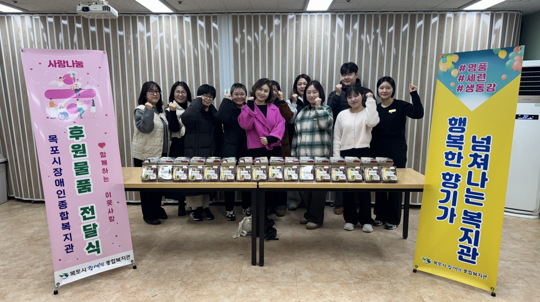 목포과학대학교 식품영양학과 관계자들이 목포시장애인종합복지관에 샌드위치 세트를 후원하고 실내에서 기념 촬영을 하고 있다. 정면 테이블 위에는 투명 용기에 포장된 샌드위치와 음료 세트가 가득 진열되어 있다. 테이블 뒤에는 11명의 관계자가 나란히 서서 오른손 주먹을 쥐고 파이팅 포즈를 취하고 있다. 좌측에는 '사랑나눔 후원 물품 전달식 목포시장애인종합복지관' 배너가, 우측에는 '넘쳐나는 복지관 행복한 향기가'라고 적힌 노란색 배너가 배치되어 있다.